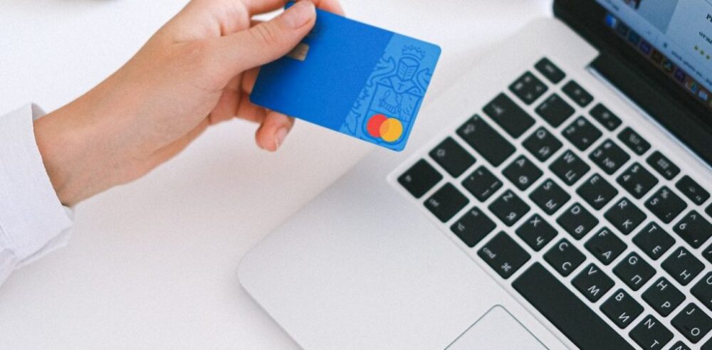 Hand holding a blue Mastercard card beside a laptop keyboard