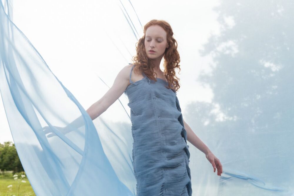 Woman wearing a pale blue dress outdoors beside flowing blue fabric
