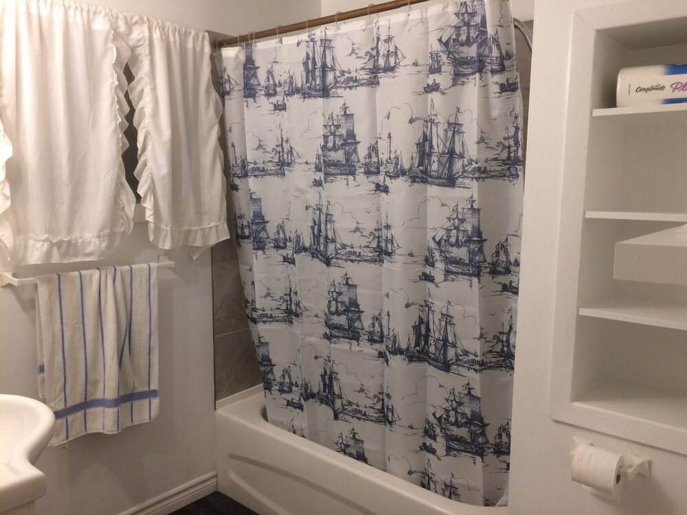 Blue-and-white sailing-ship print shower curtain hanging beside a white window curtain and striped towel
