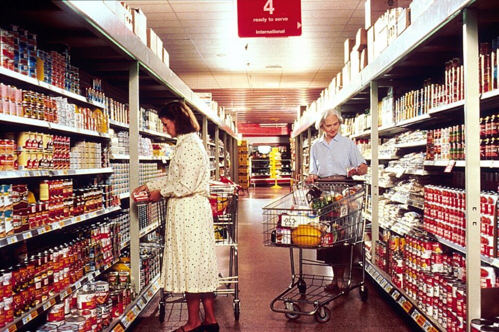 Woman shopping in a grocery store aisle with a cart full of items, illustrating bulk and multi-item purchasing decisions.