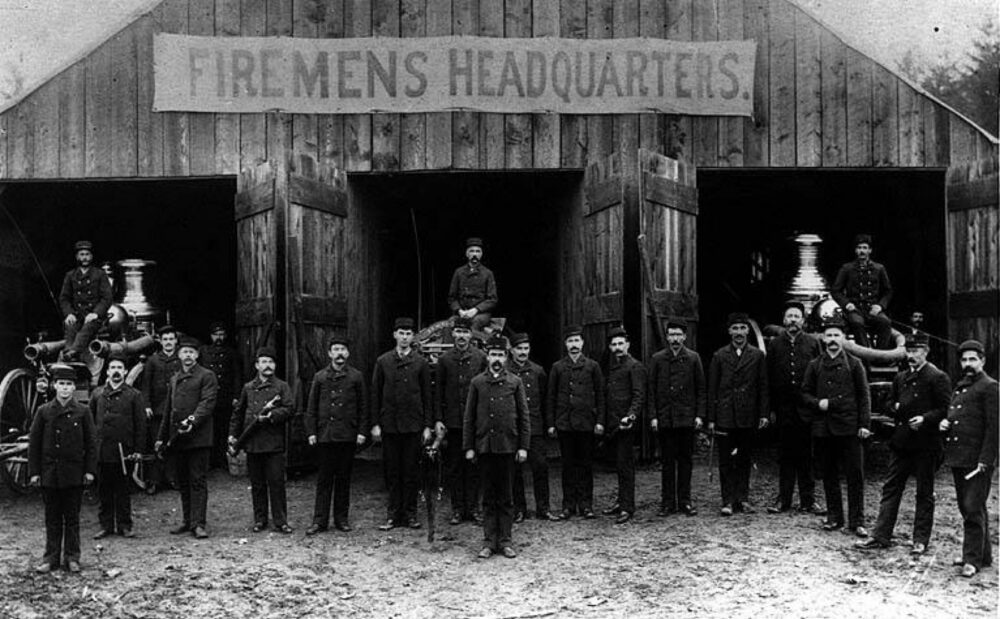 Historic image of firemen outside a wooden building with a temporary banner over its doors, symbolising temporary access solutions