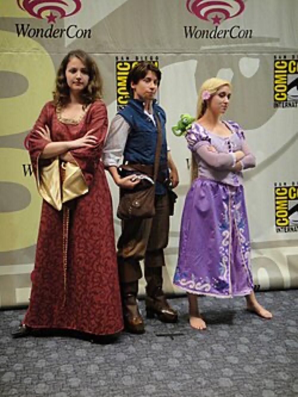 Cosplayers dressed as Mother Gothel, Flynn Rider and Rapunzel on stage at a convention, illustrating the detail level possible in Tangled costumes.