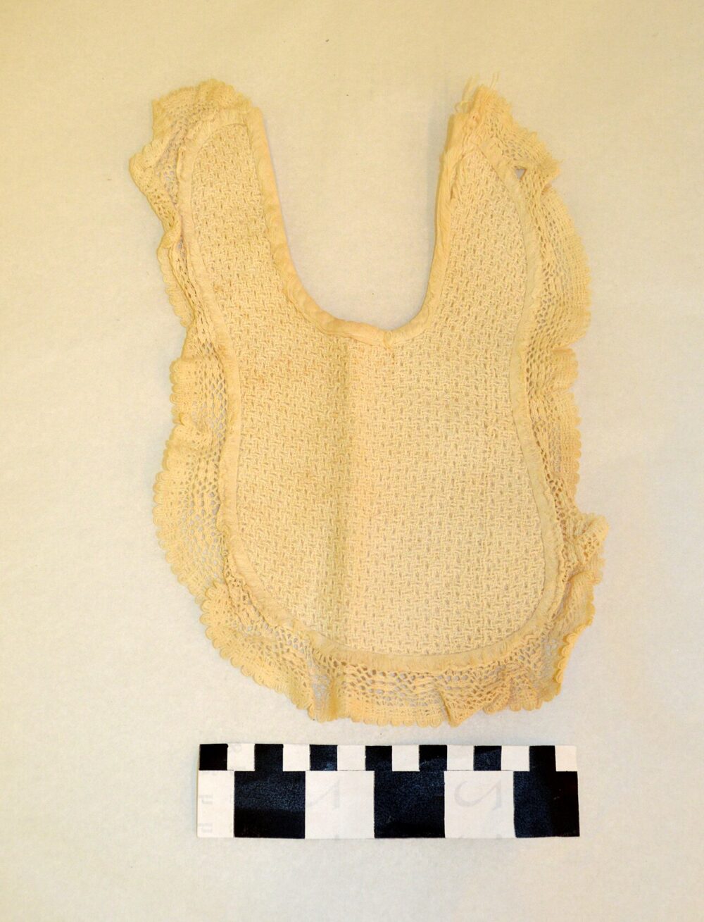 Cream-colored historical baby bib photographed flat with a measurement scale