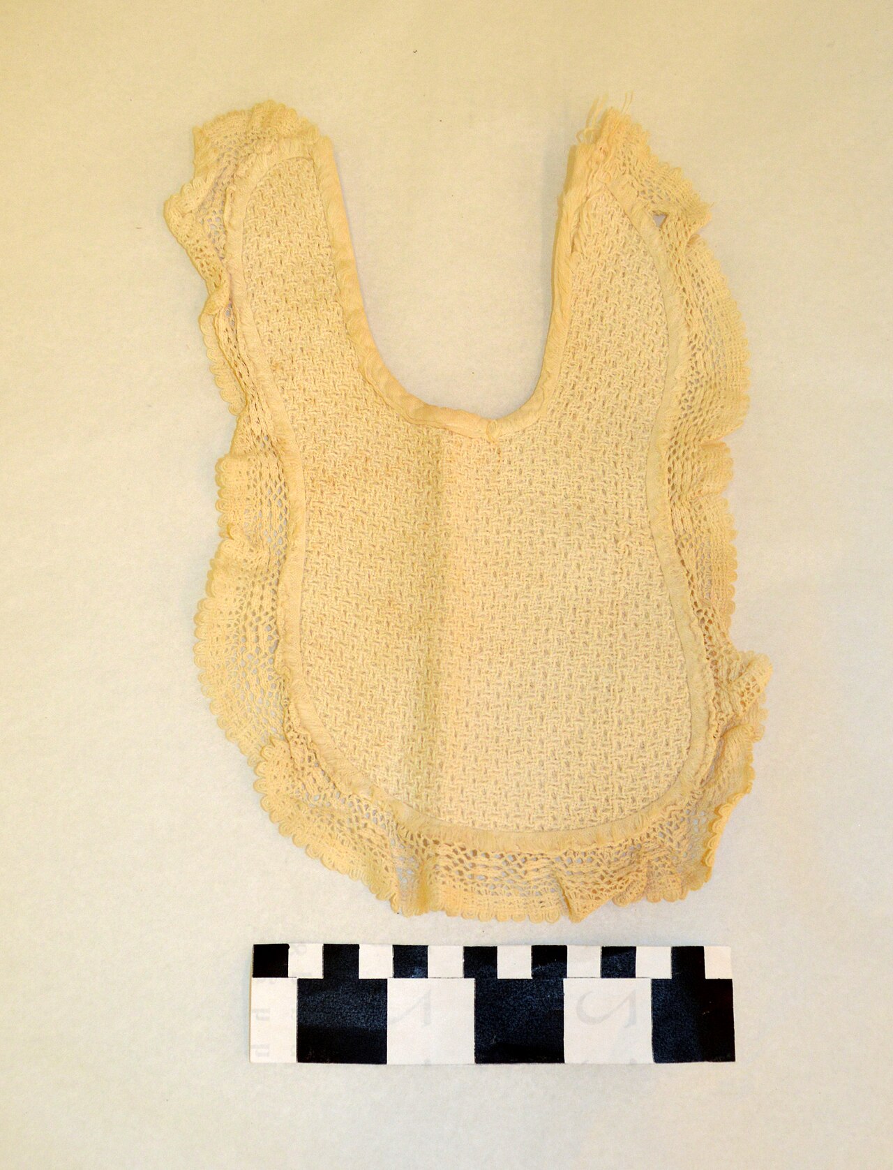 Cream-colored historical baby bib photographed flat with a measurement scale